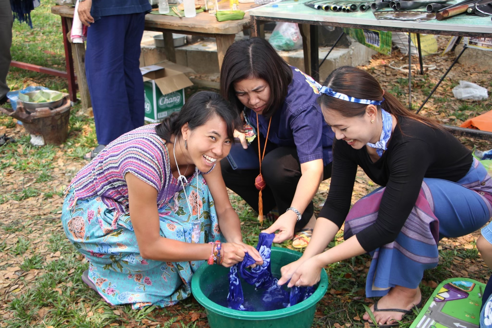 Three woman gather around a bucket filled with water, squeezing water out of fabric. The woman on the left smiles at the camera, while the woman in the center and the woman on the right are focused on their task. They are outside on the grass. 