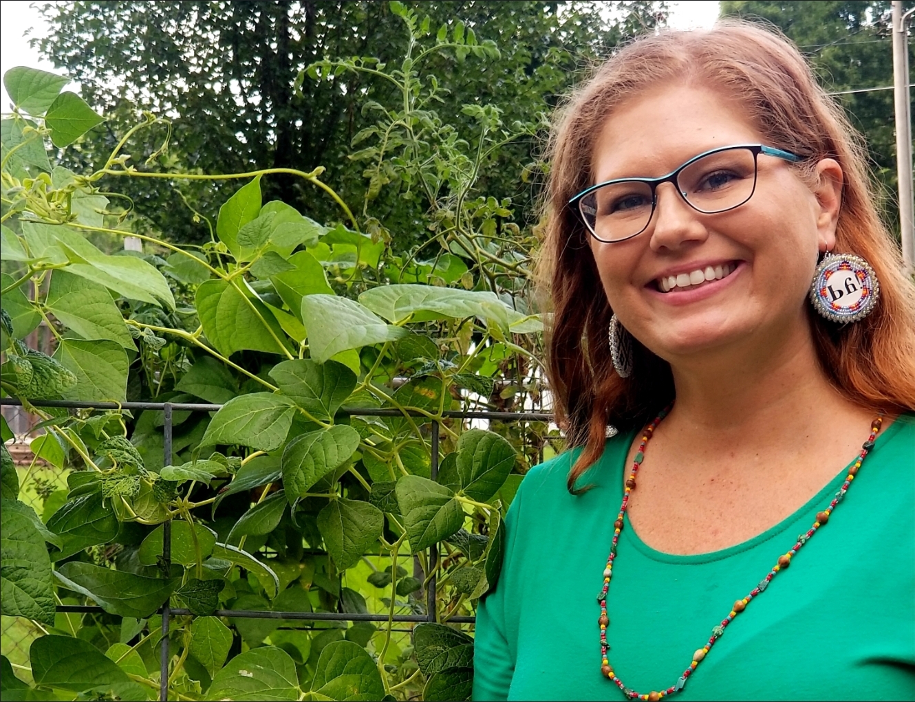 A woman wearing glasses and round dangly earrings stands at the right of the picture next to plants. She smiles at the camera.