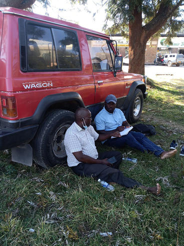 Nyachega sitting on the ground with another man, leaning against a red vehicle