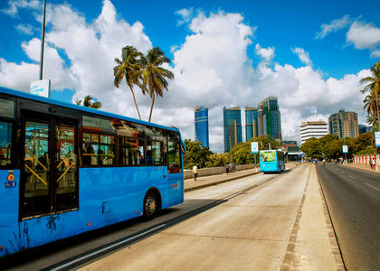 A bus is pictured on the left, traveling toward a city with skyscrapers and palm trees ahead. The sky is bright and sunny, with white puffy clouds indicating warm weather.