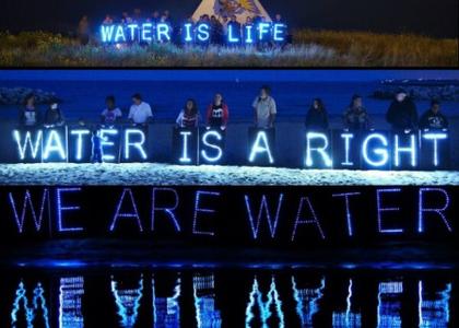 A group of people stand by water at night, holding neon letters that spell out "Water is Life" and, closer to the water, another group of people hold neon letters that spell out "Water is a Right." The neon letters reflect in the water. 