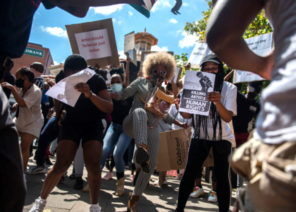 Several people hold signs in an apparent protest. A young woman with natural, Afro-styled hair kicks the air in the center of the image.